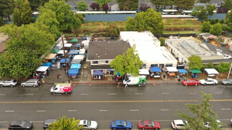 Aerial view of a street market with colorful tents lining the sidewalks, people browsing booths, and vehicles parked along a wet road.