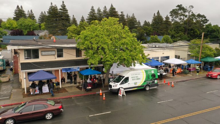 Street-level view of a community health fair with multiple tents and booths along a sidewalk, people browsing and talking under canopies, a white health services van parked nearby.