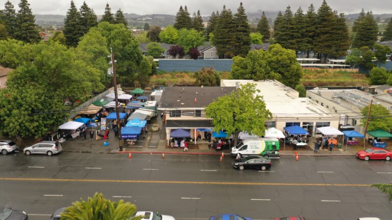 Aerial view of a busy street market with colorful tents, stalls, and shoppers along a storefront row.