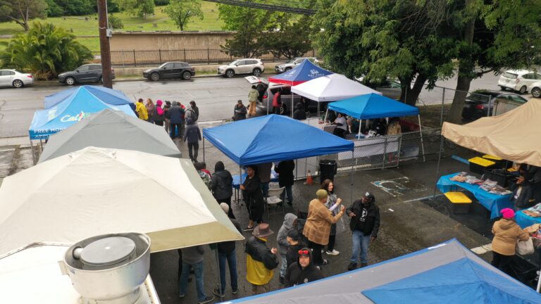 Outdoor street market or community event with multiple blue and white tents and people mingling along a wet road fence in the background.