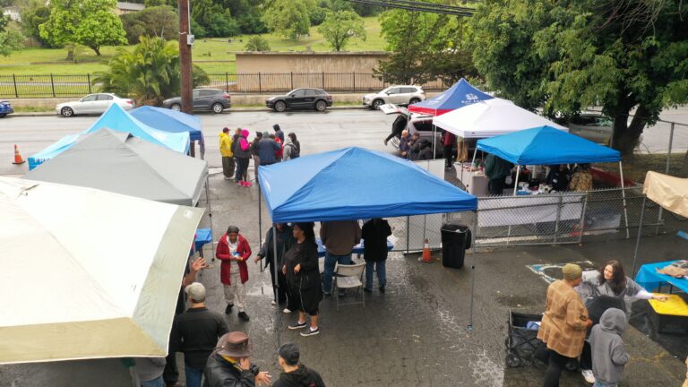 Outdoor event in a parking lot with multiple blue and white tents and people gathered around a central area under cloudy skies.
