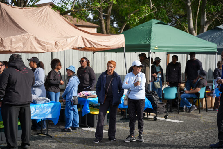 Group of people under beige and green canopies at an outdoor event, chatting around blue-tablecloth tables with supplies nearby.