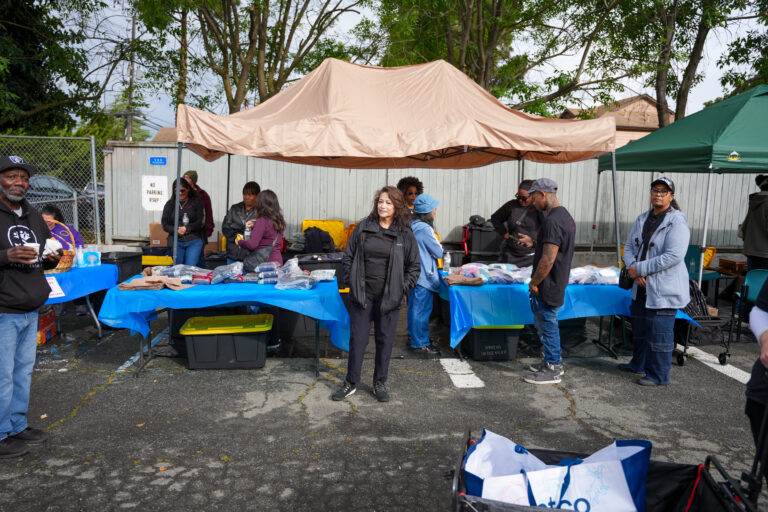People under a large beige canopy browsing tables of donated clothes and goods at an outdoor market/event.