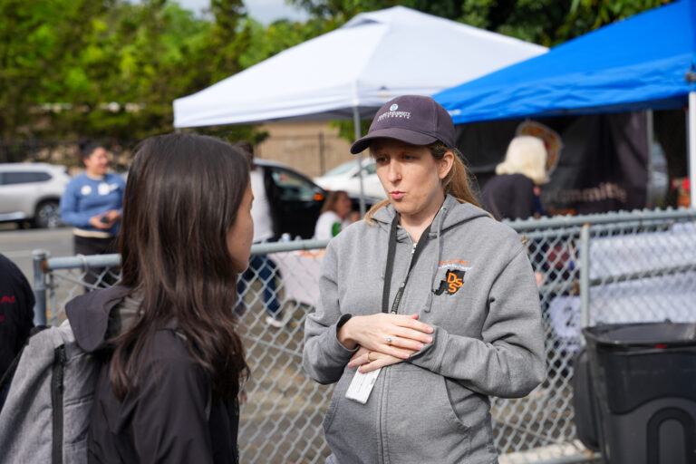Woman in a gray hoodie with a cap talking to another person at an outdoor event, with white and blue tents in the background behind a chain-link fence.