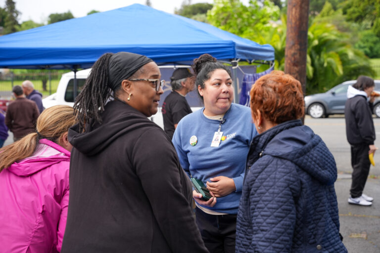 Group of people talking outdoors under a blue canopy tent at an event, wearing jackets and badges visible on a staff member's blue shirt.