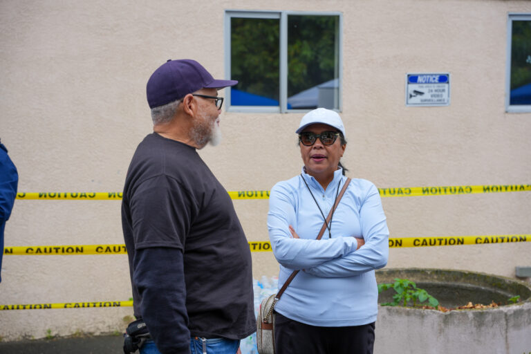 Two people talking beside a beige building behind yellow caution tape and windows in the background.