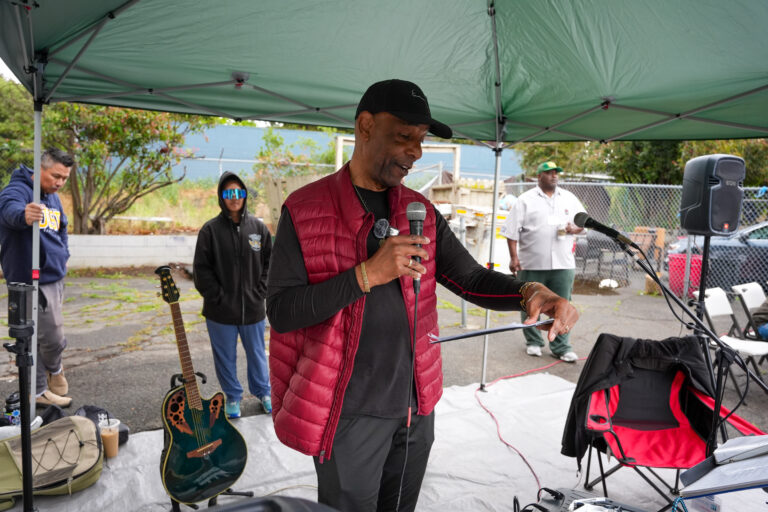 Man in a red vest speaks into a handheld microphone under a green canopy, with a guitar and audio gear nearby outdoors.