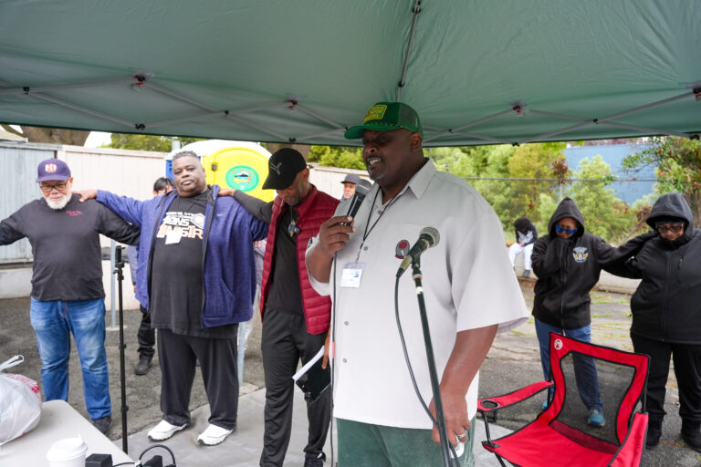 A man in a white shirt speaks into a microphone under a teal canopy while others stand with arms around each other at an outdoor gathering.