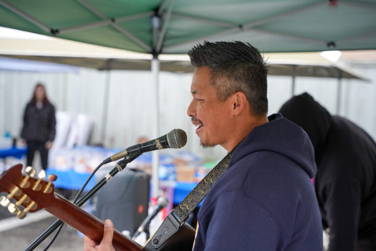 Man singing into a microphone while playing a guitar under a green canopy outdoors.