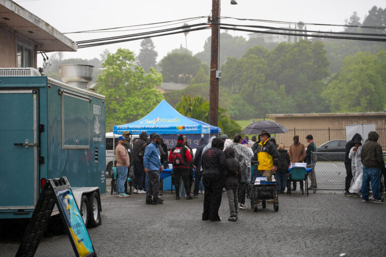 Crowd gathered in rainy parking lot around a blue CalFresh tent for a food distribution; a large teal service trailer is on the left.