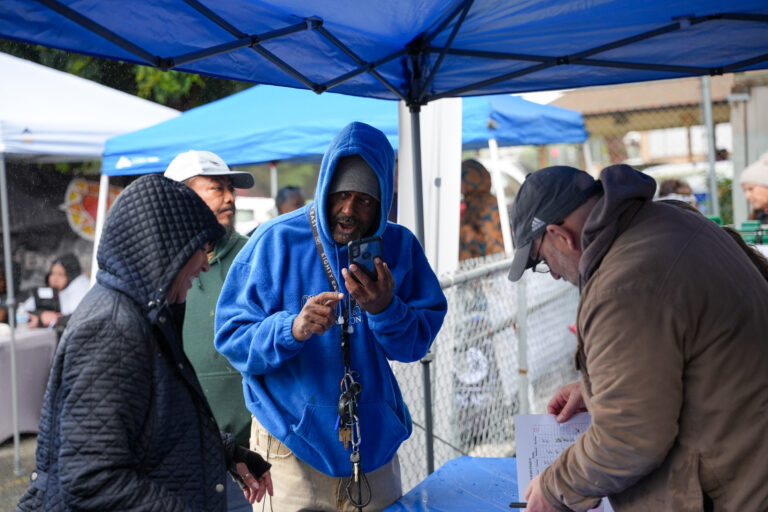 Man in a blue hoodie speaks and gestures to two others under blue canopies at an outdoor event.