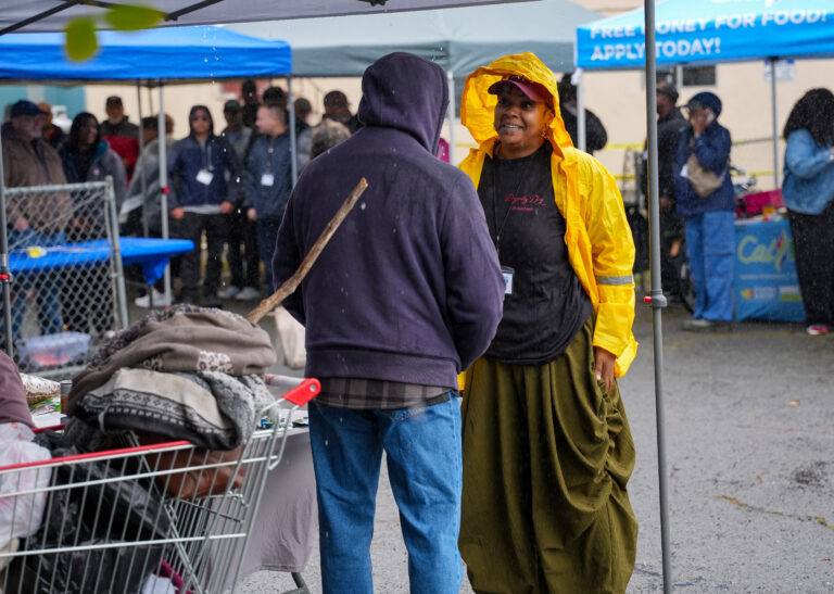 Volunteer in a bright yellow raincoat speaks with a person in a dark hoodie at a rain-soaked clothing donation booth, with a cart of clothes nearby.