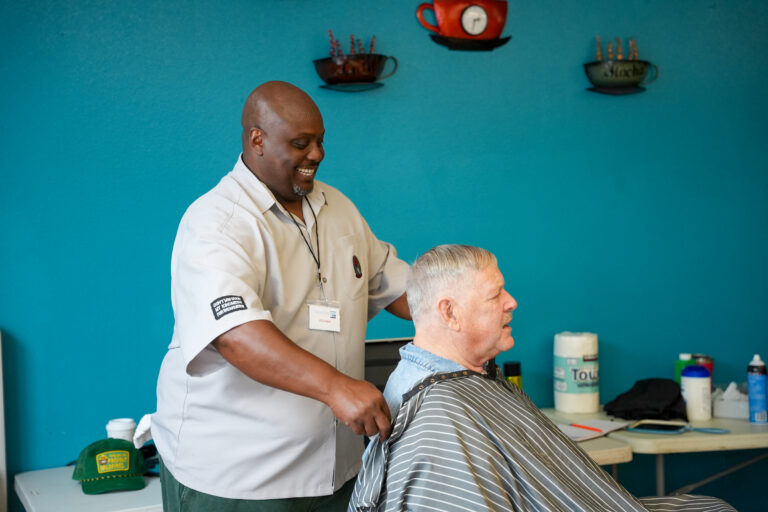 Barber in a white shirt trims an elderly man's hair in a blue-walled shop, both smiling.
