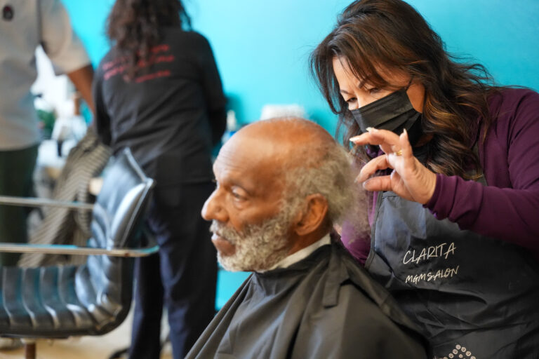 Hairdresser wearing a black mask shapes an elderly man's hair in a salon with a bright turquoise wall behind them.