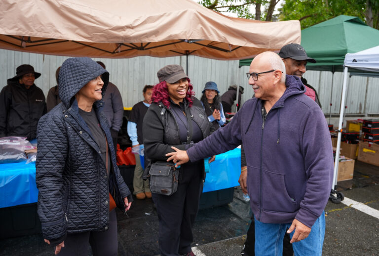 People greet each other at an outdoor market under beige tents, smiling and chatting while a man in a purple hoodie shakes hands with a woman in a quilted jacket.