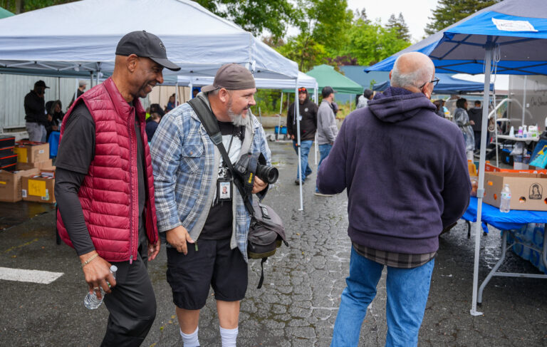 Two men chat and laugh at an outdoor market with vendor tents and boxes in the background, one with a camera.