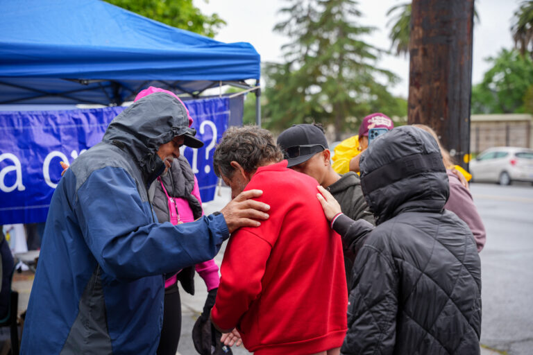 Group of people wearing jackets, hugging a person in a red hoodie near a blue tent. Outdoor event.