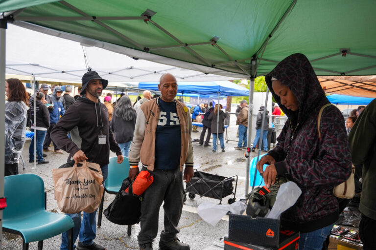 People shopping under green market tents; a man in a tan jacket stands center with bags, others browse stalls in the background.
