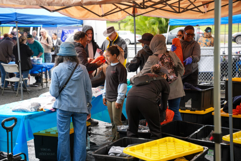 People under canopy at a clothing drive, handing out garments into blue tables and yellow bins.
