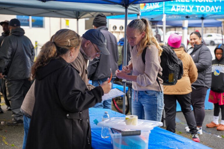 Man in a cap signs a clipboard at a table under blue tents while a woman with a backpack speaks to him at an outdoor event.