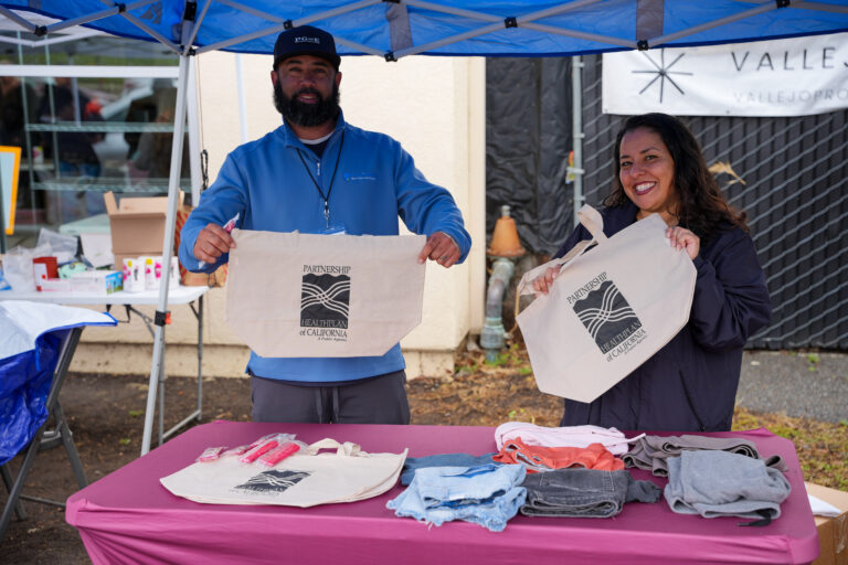 Two volunteers at an outdoor booth handing out beige tote bags with the Health Plan of California logo.