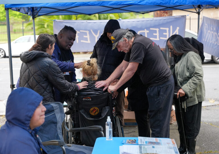 Group of people under a blue canopy helping a person in a wheelchair during an outdoor event, with pamphlets on a table nearby and a banner in the background.